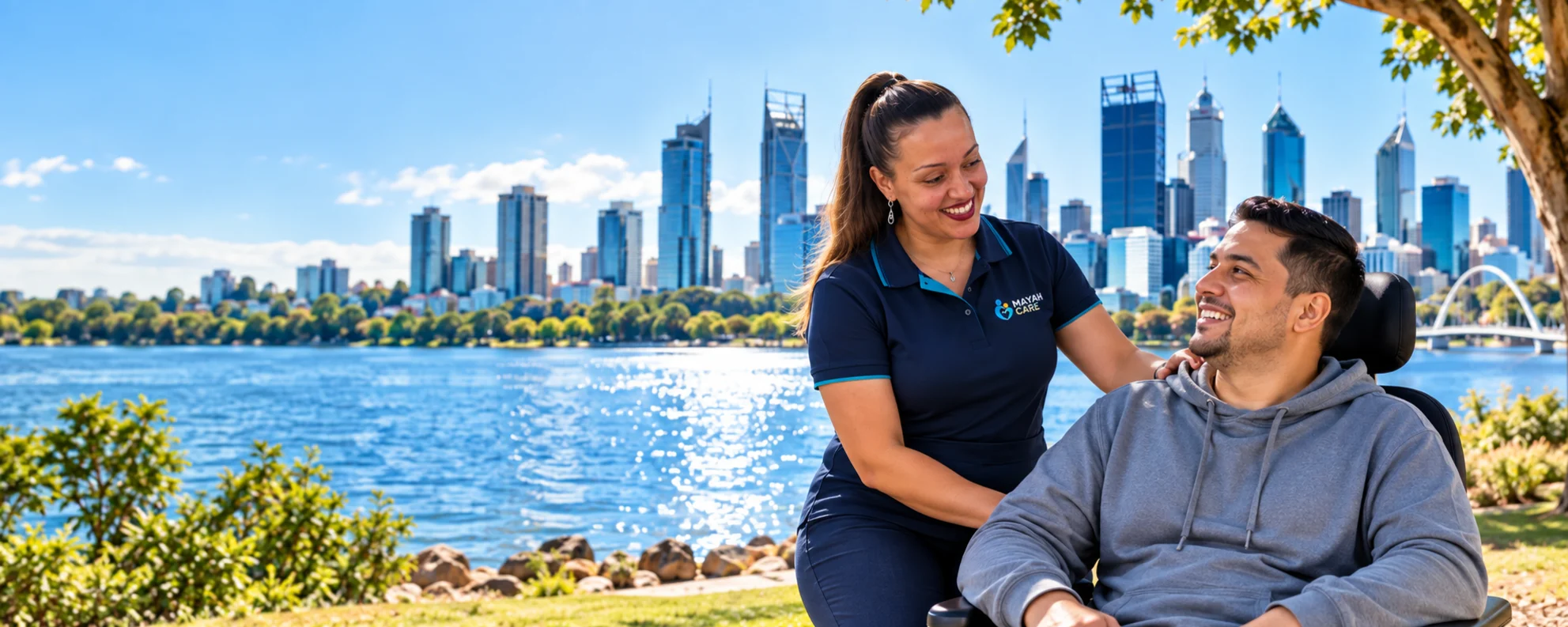 A support worker with her hand on the shoulder of a smiling man in a power wheelchair, on a riverside lawn with a city skyline behind
